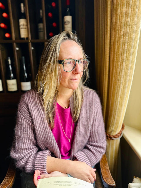 Woman wearing glasses and a purple cardigan in a room with wine bottles in the background
